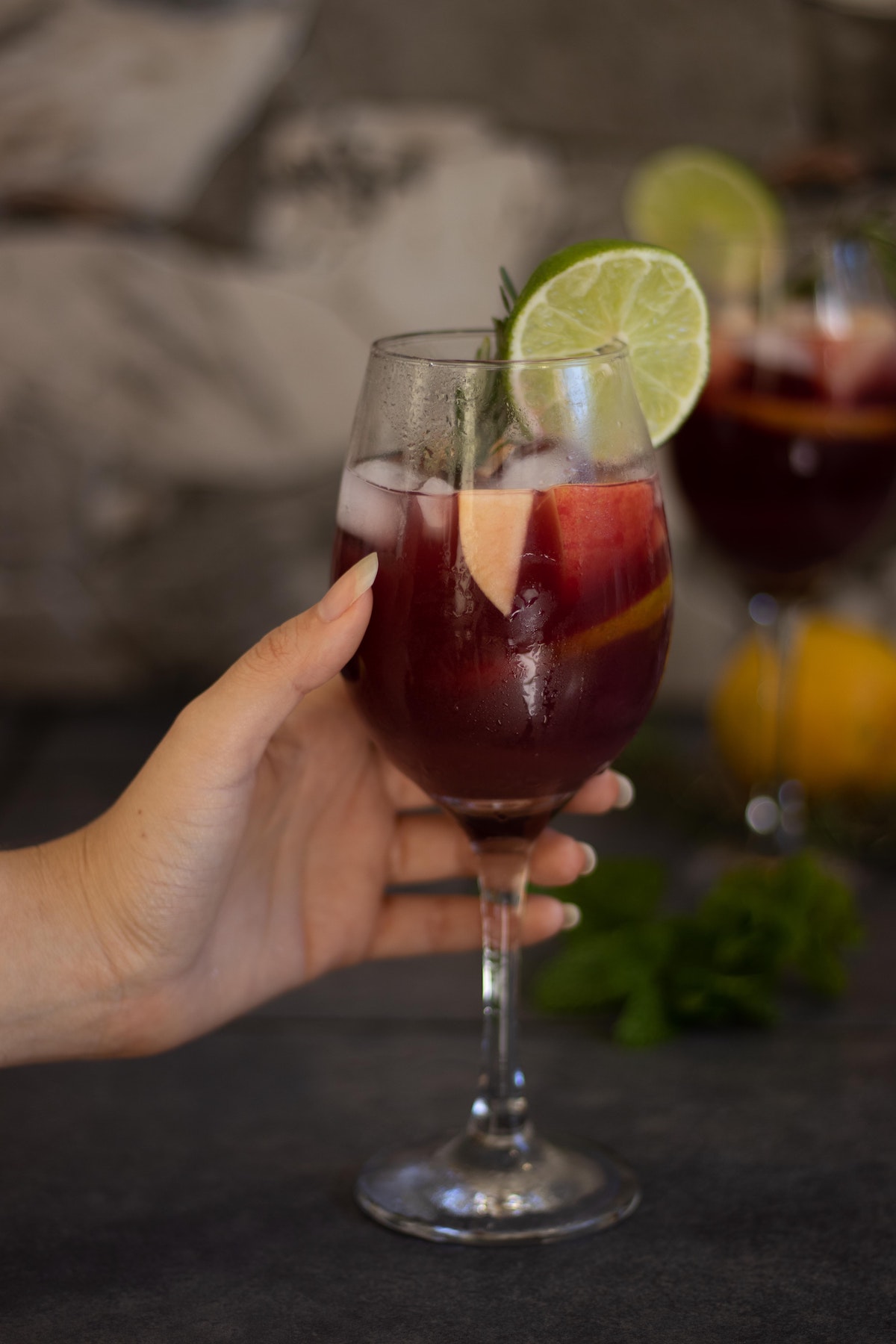 Person's hand holding a wine glass of sangria garnished with fruit and fresh herbs