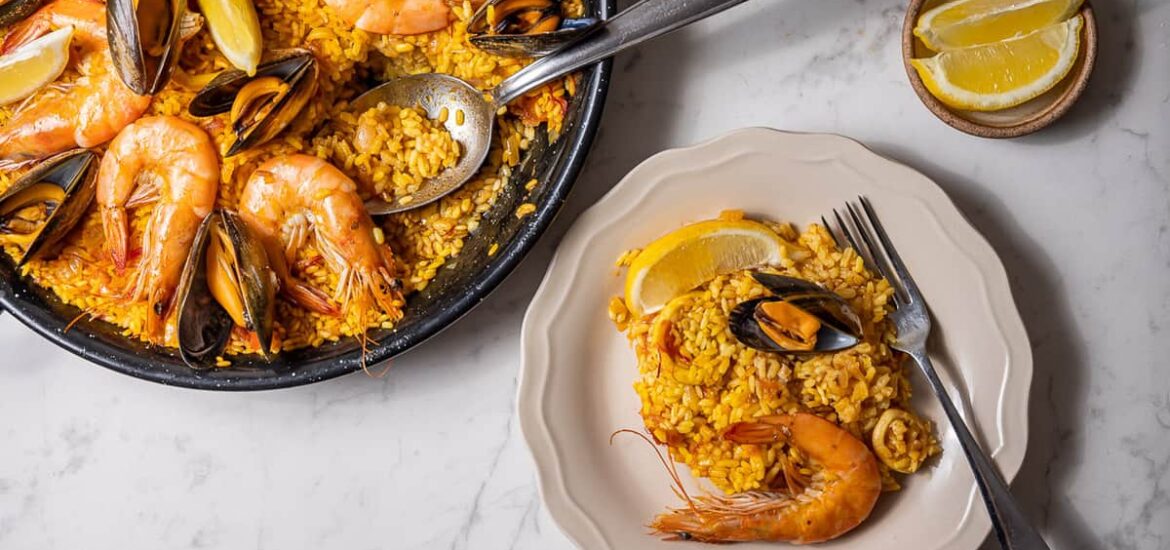 Overhead shot of a pan of seafood paella with a small amount served out onto a white plate.