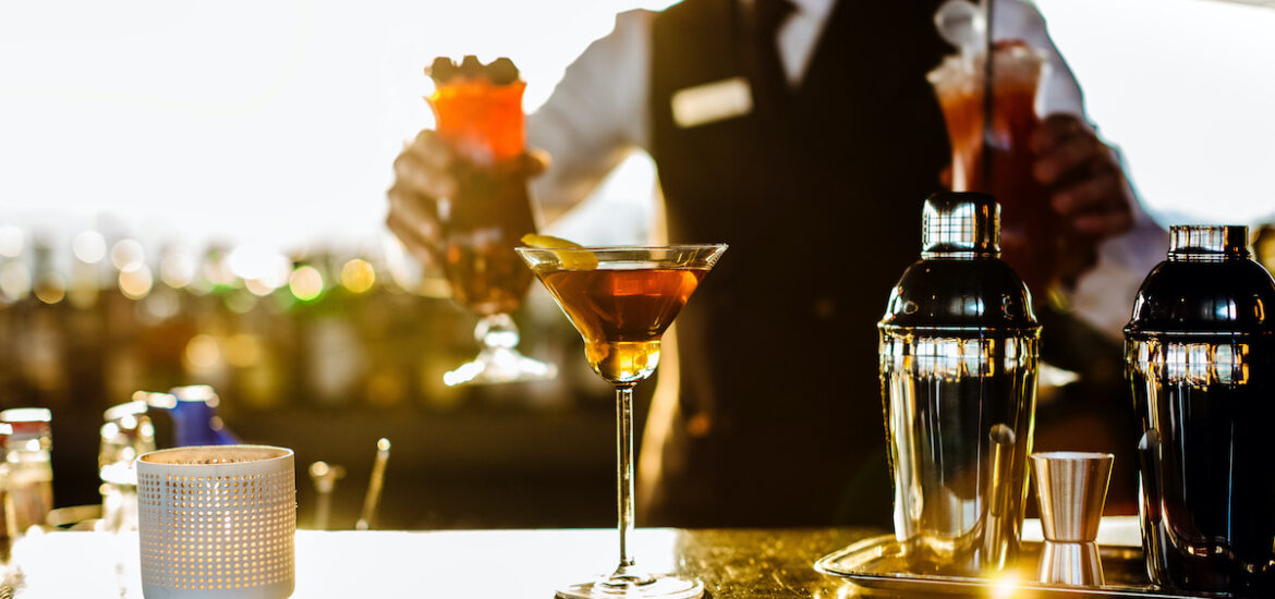 Brown cocktail in a martini glass at an outdoor terrace with a uniformed bartender in the background