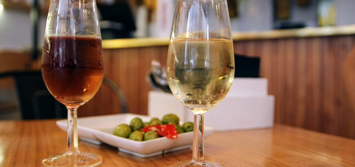 Two glasses of sherry wine (one amber-colored and one light yellow) in front of a dish of olives on a wooden tabletop.