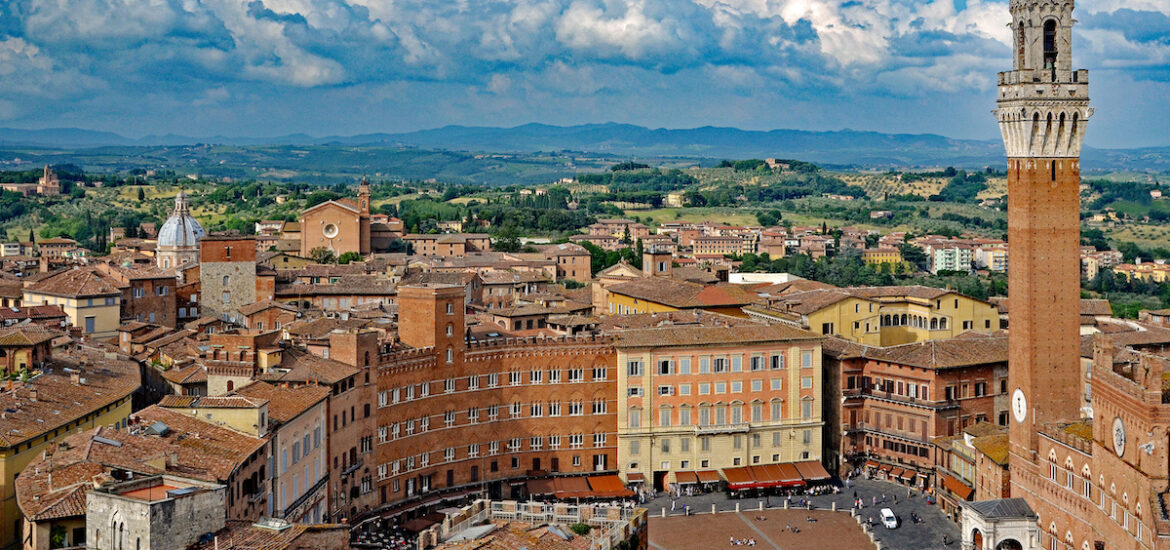 Aerial view of Siena, Italy, with brown historic buildings and a prominent tower at front right, with green countryside in the background