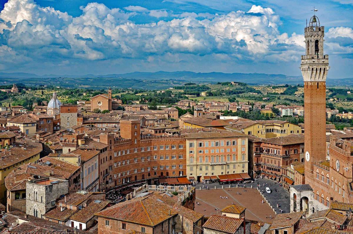 Aerial view of Siena, Italy, with brown historic buildings and a prominent tower at front right, with green countryside in the background