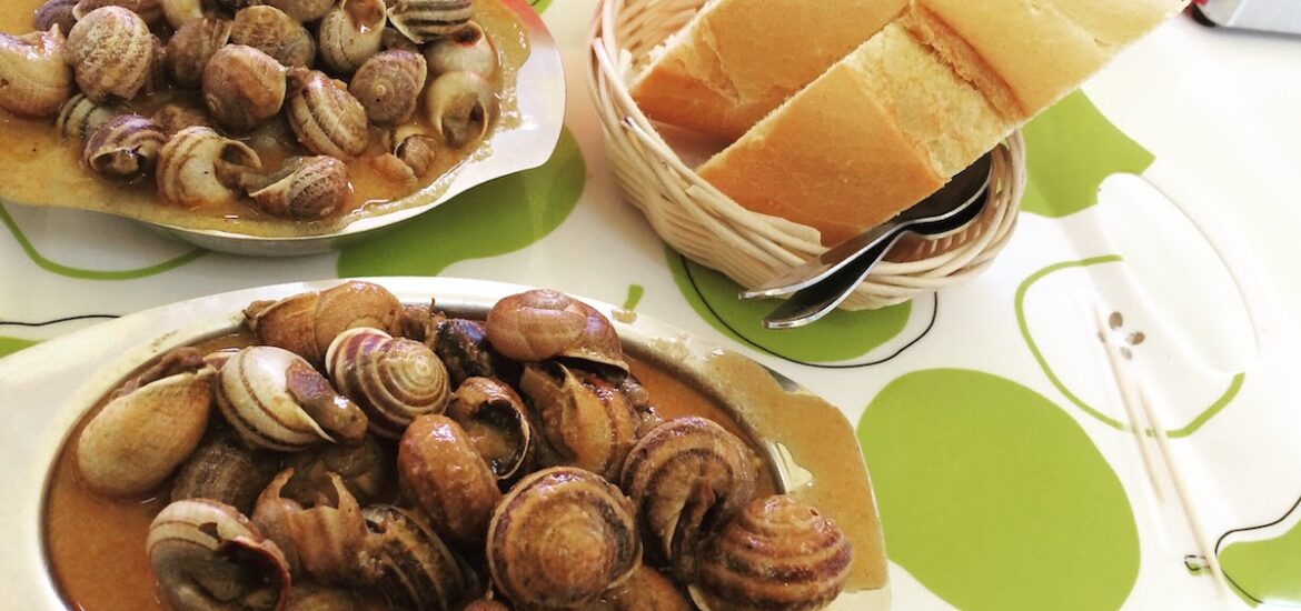 Two small metal plates of snails in broth next to a basket of bread.