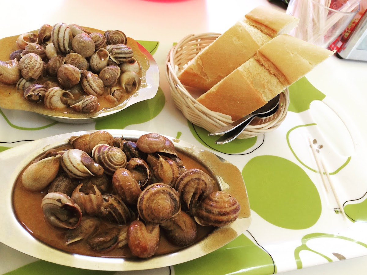 Two small metal plates of snails in broth next to a basket of bread.