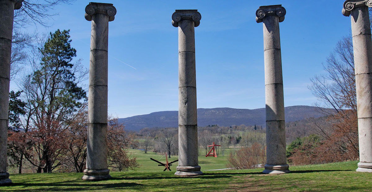 Five large stone columns in an open outdoor space with several other sculptures visible in the background