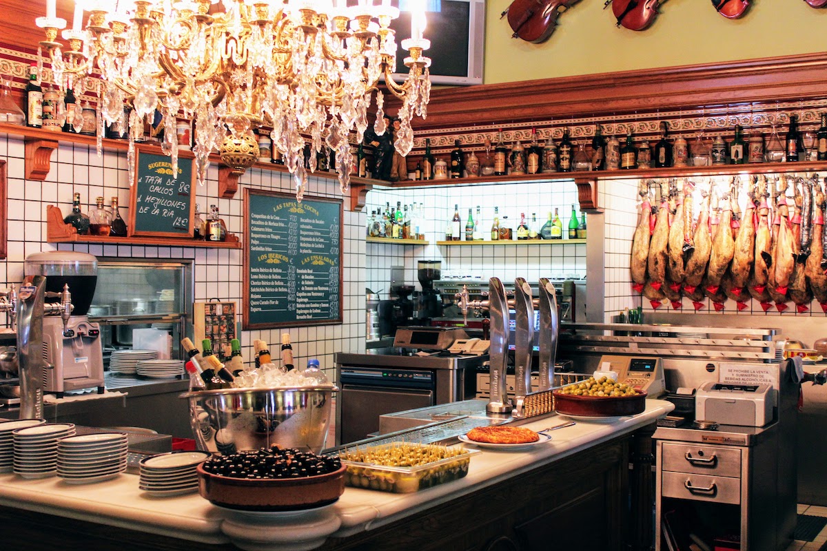 Interior of a typical Spanish bar with large pots of olives on the bar top and a large crystal chandelier overhead.