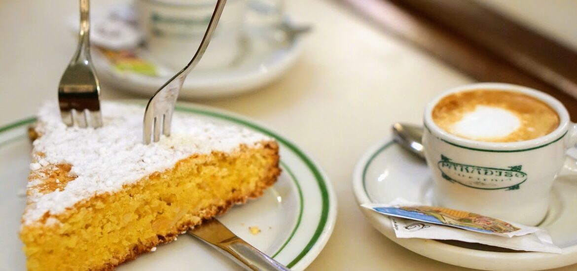 A slice of almond cake dusted with powdered sugar on a table beside two cups of coffee.