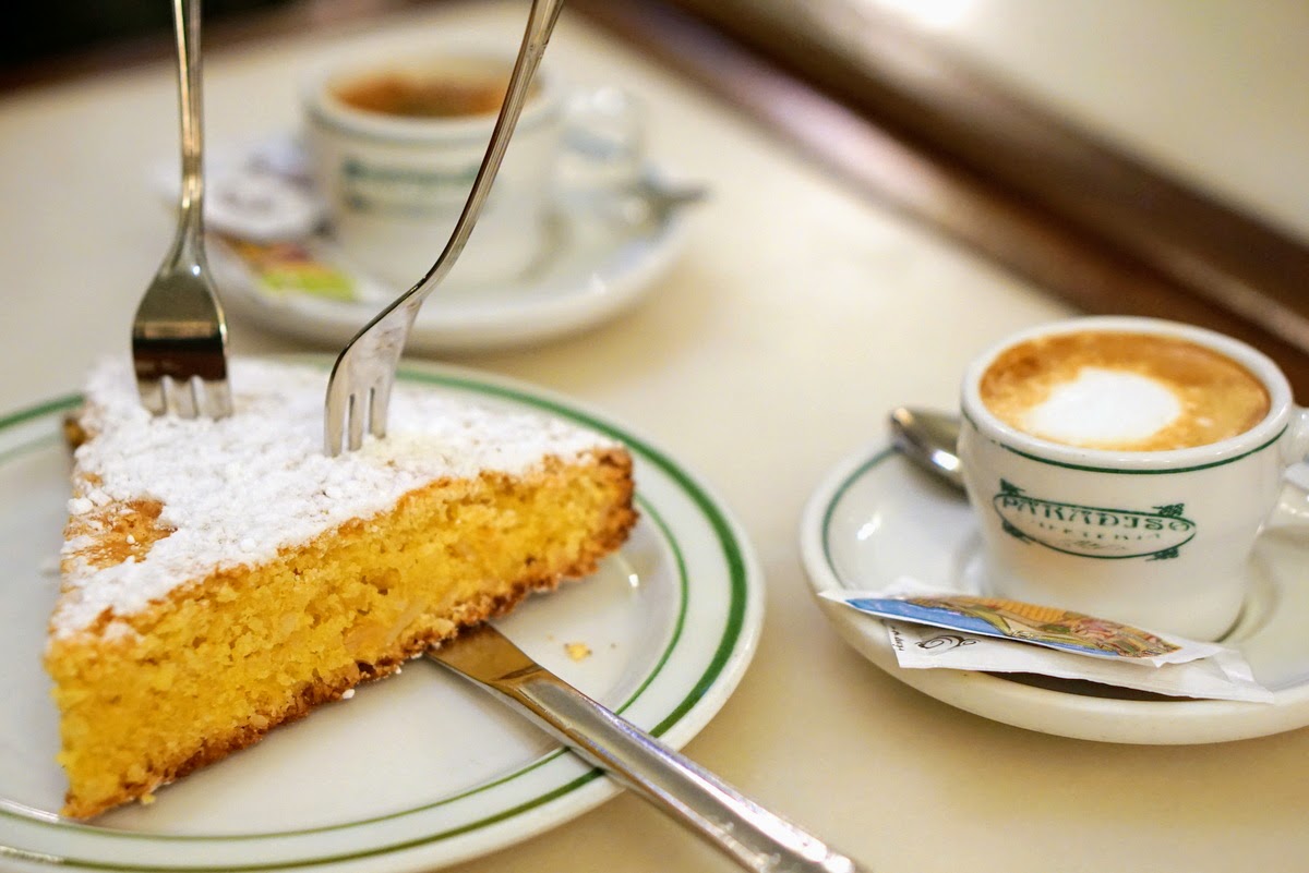 A slice of Galician almond cake dusted with powdered sugar on a table beside two cups of coffee.