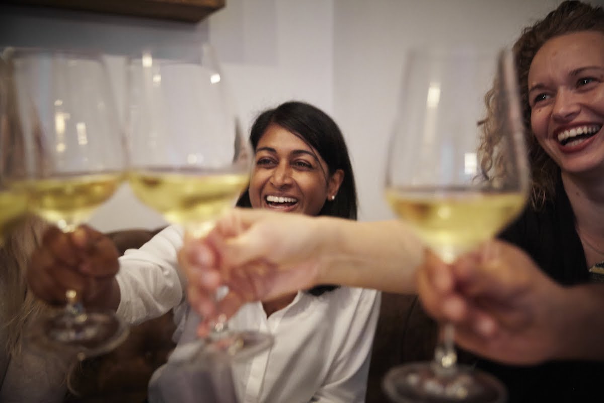 Two women smiling while toasting with glasses of white wine.