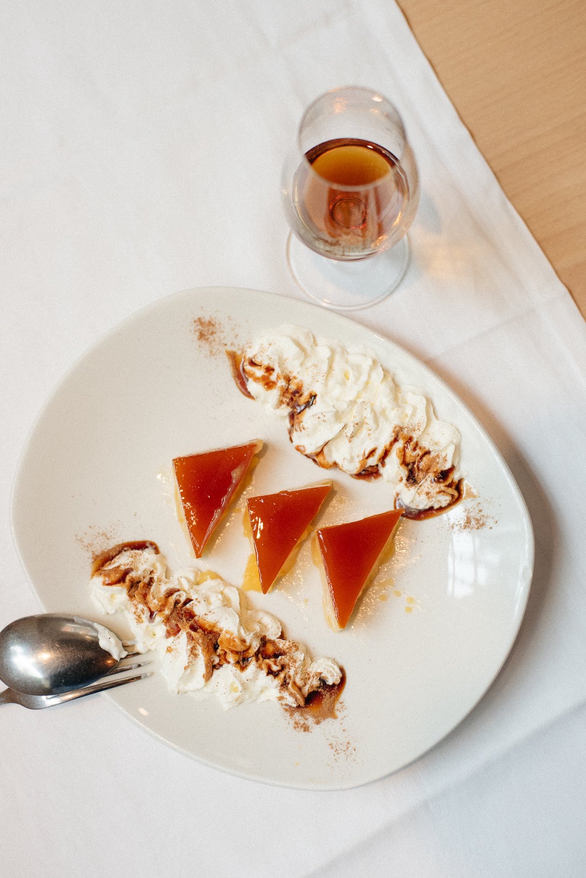 Three triangle-shaped pieces of tocino de cielo, a Spanish dessert, on a plate decorated with whipped cream beside a glass of brown dessert wine.