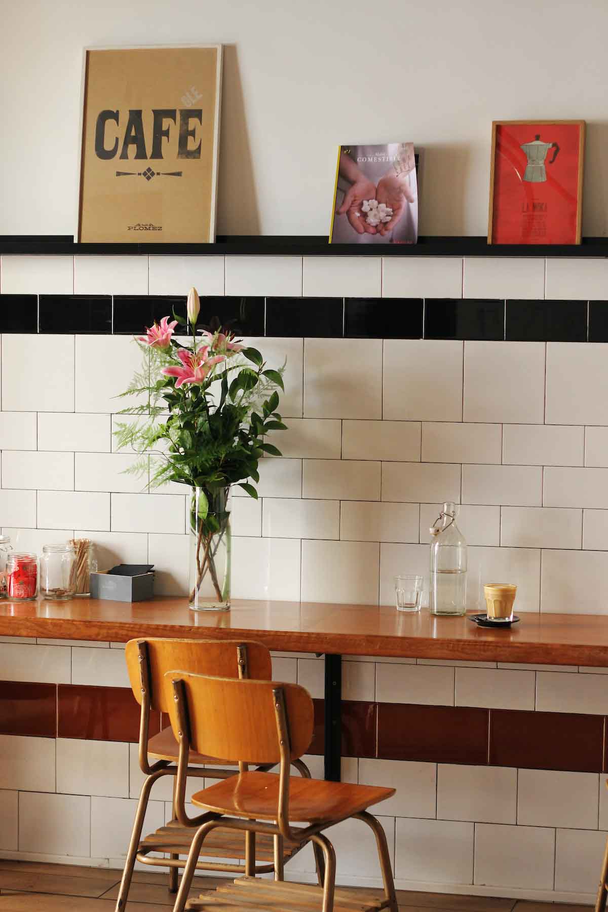 Wooden counter against a wall with two wooden chairs in front and a cup of coffee on top, with coffee-themed decor on the above wall.