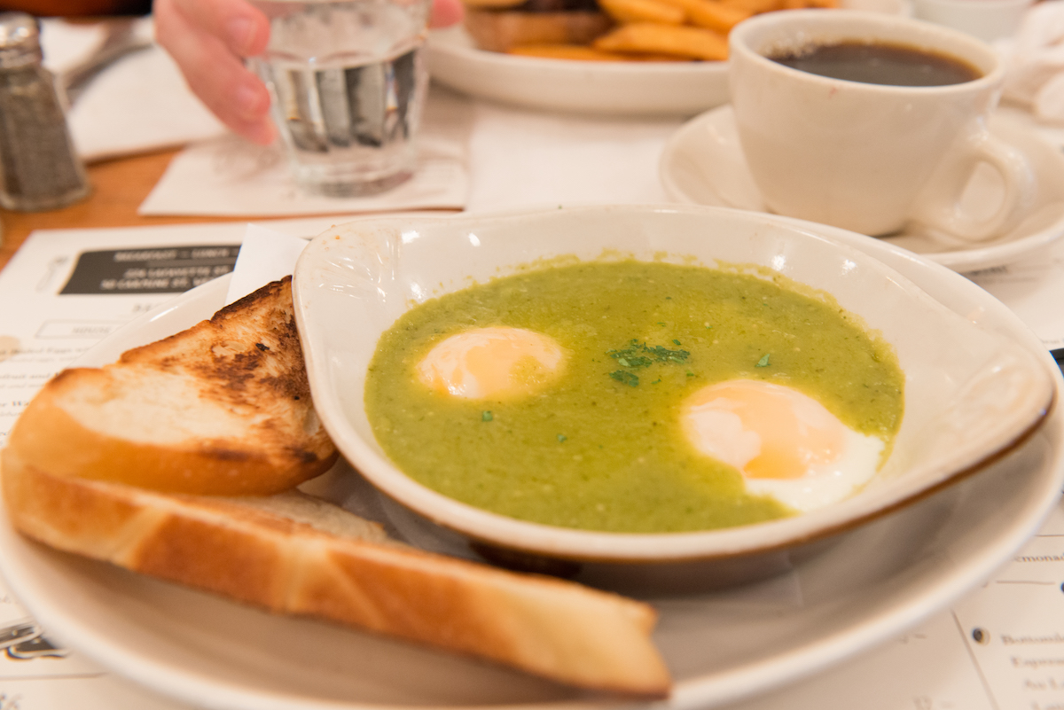 Cooked eggs in a green sauce in a dish on a white plate beside two pieces of toast