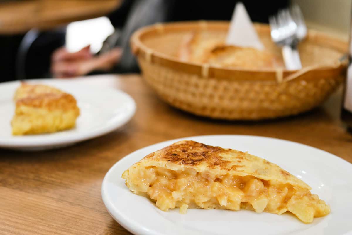 Two slices of Spanish potato omelet on separate white plates with a brown bread basket in the background.