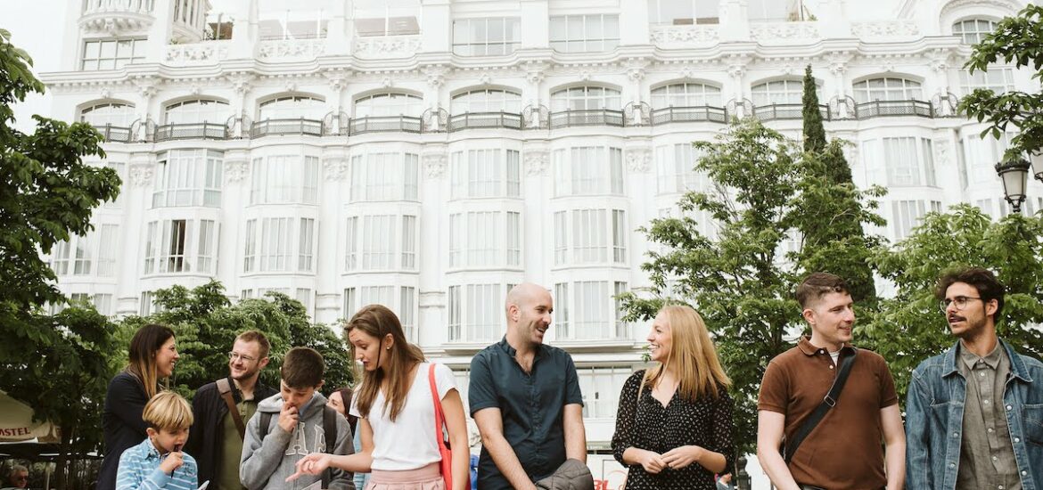 7 adults and 2 young boys standing in a leafy green plaza in front of a large white hotel.