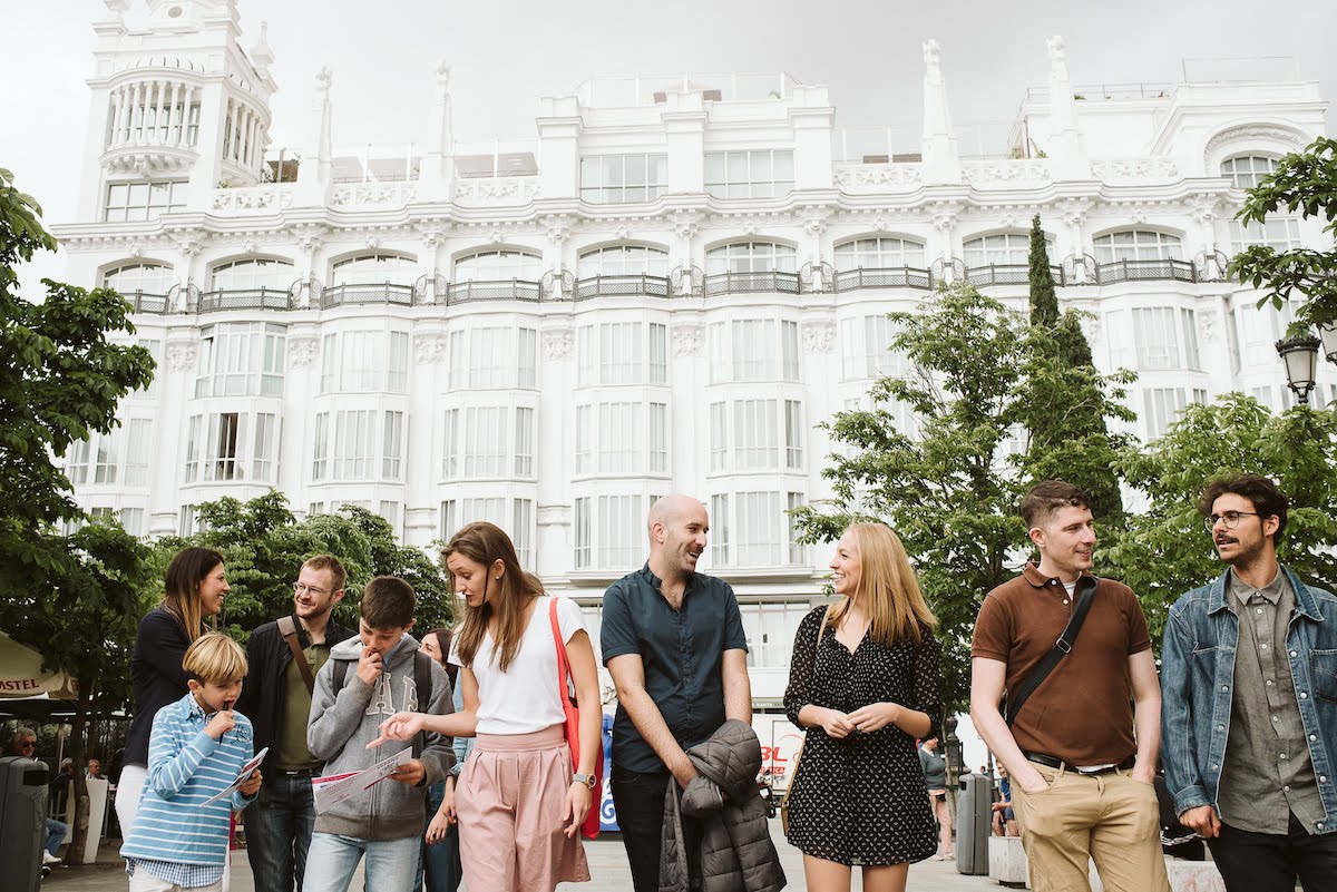 7 adults and 2 young boys standing in a plaza in front of several trees and a large white hotel.