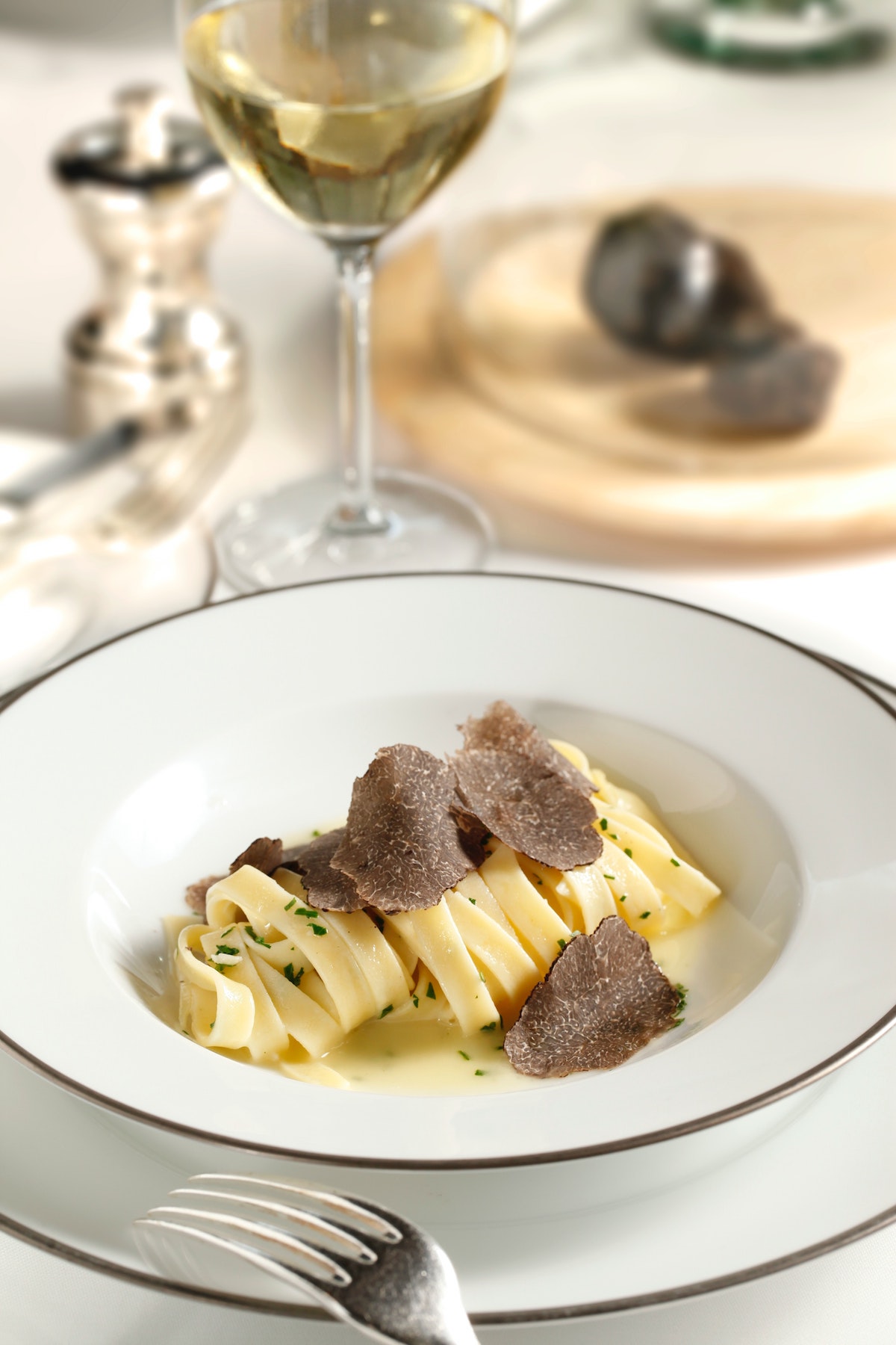 Pasta dish made with wide, flat noodles and topped with truffle shavings on a table beside a glass of white wine.