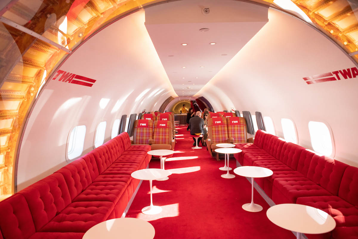 Restaurant inside a refurbished TWA airplane, with red carpeting and bench seating, small round white tables, and plaid airplane seats in the background
