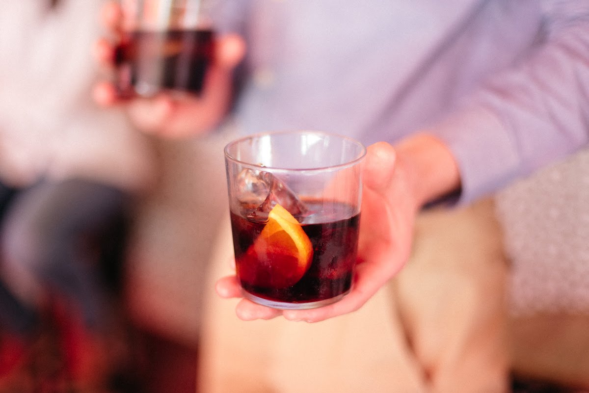 Close up of a person's hand holding a vermouth glass with an orange slice garnish