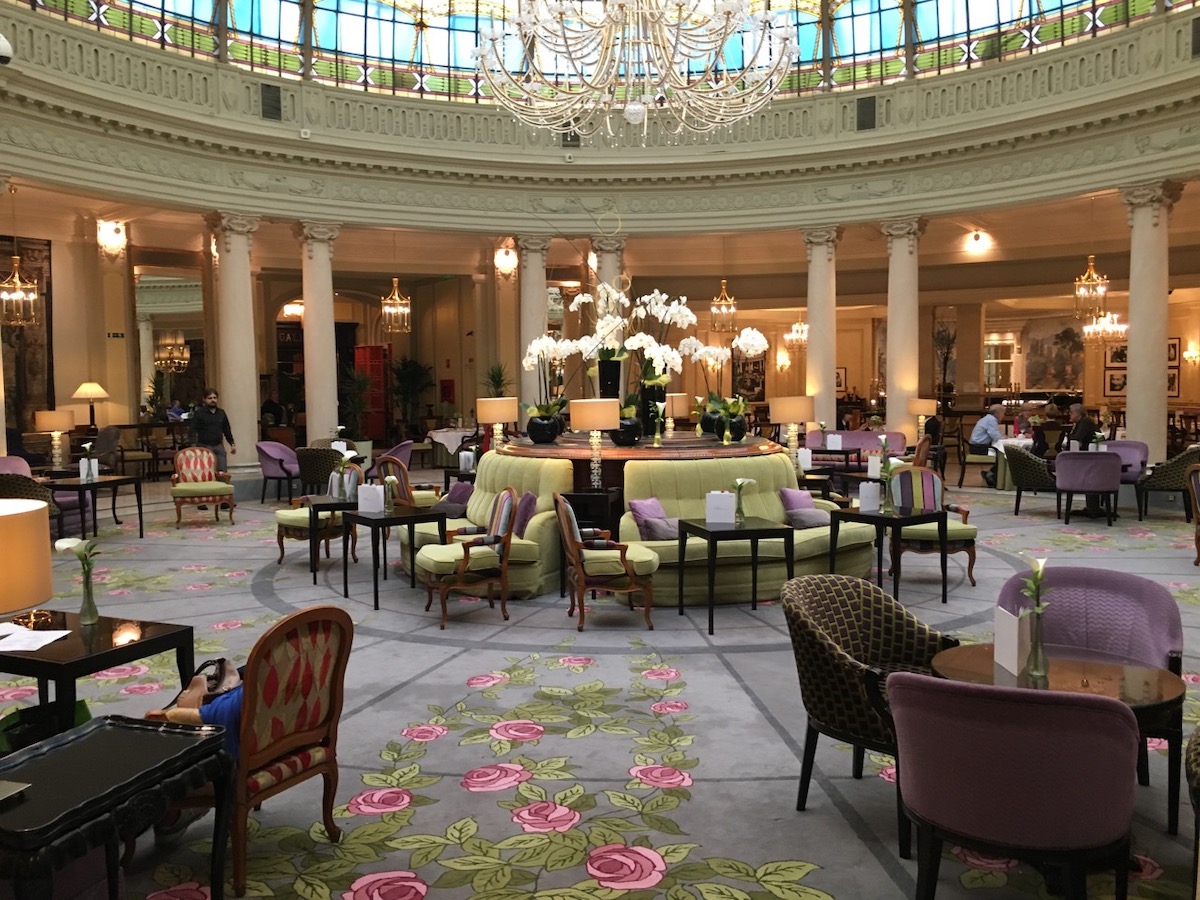 Spacious hotel restaurant under a large glass dome and crystal chandelier, with light green and purple decor.
