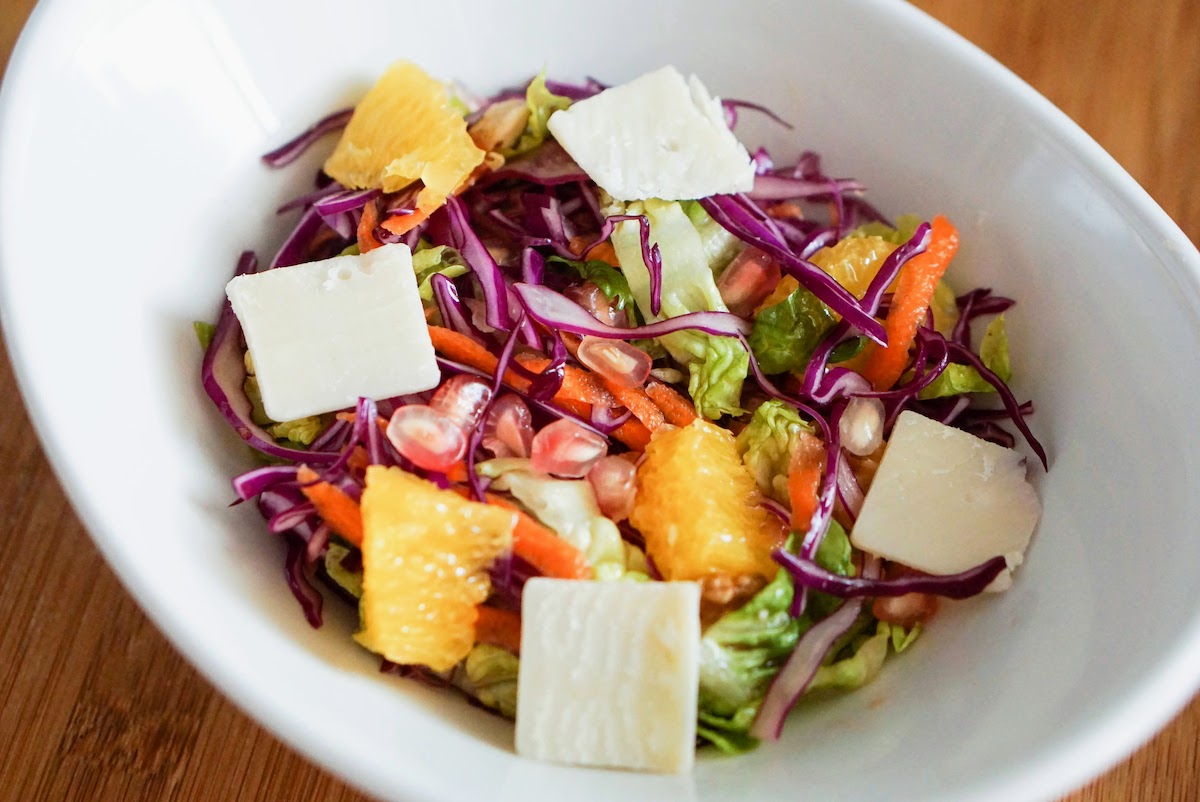 Salad with orange, pomegranate, red cabbage, lettuce, and shaved Manchego cheese in a white bowl